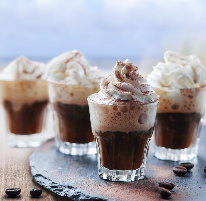 Vier kleine Gläser mit schaumigem Kaffee, gekrönt von Sahne und Schokoladenstreuseln, stehen auf einem rustikalen Tisch.