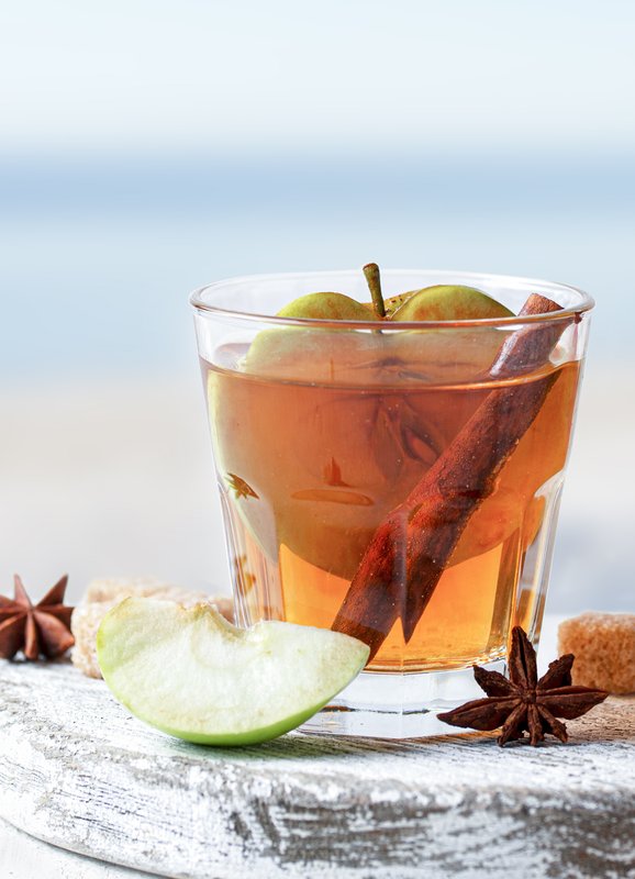 Ein Glas mit einem warmen Getränk, gefüllt mit Äpfeln und Zimt, steht auf einem rustikalen Holzbrett neben frischen Äpfeln.