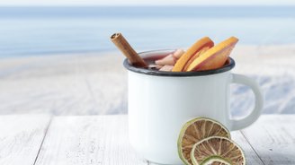 Ein weißer Becher mit Zimtstange und frischen Obstscheiben steht auf einem Holztisch, im Hintergrund der ruhige Strand.