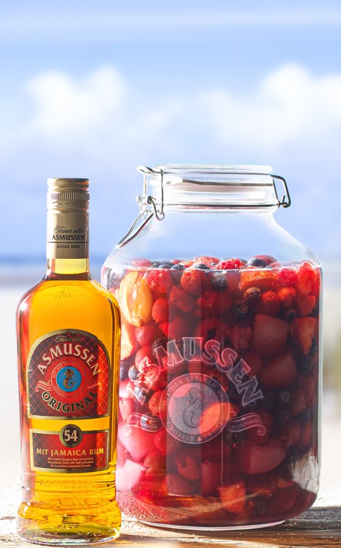Eine Flasche Asmussen Rum steht neben einem großen Glas mit bunten Früchten, vor einer malerischen Strandkulisse.