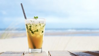 Ein erfrischendes Getränk mit Minze und Eiswürfeln steht auf einem Holztisch, während der Strand im Hintergrund verschwommen ist.