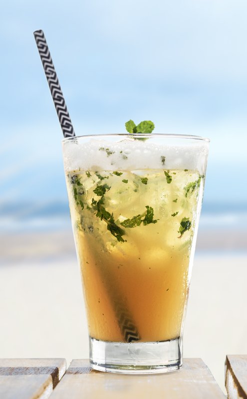 Ein erfrischendes Getränk mit Minze und Eiswürfeln steht auf einem Holztisch, während der Strand im Hintergrund verschwommen ist.