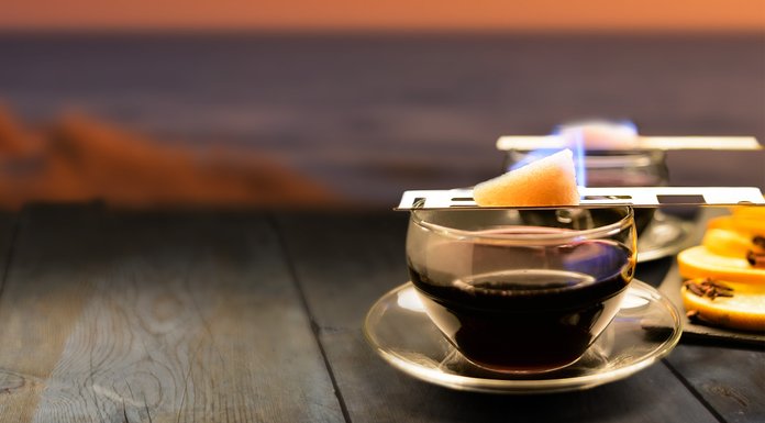 Ein Glas mit heißem Getränk steht auf einem Tisch, während ein Stück Zucker flambiert wird und orangefarbene Scheiben daneben liegen.