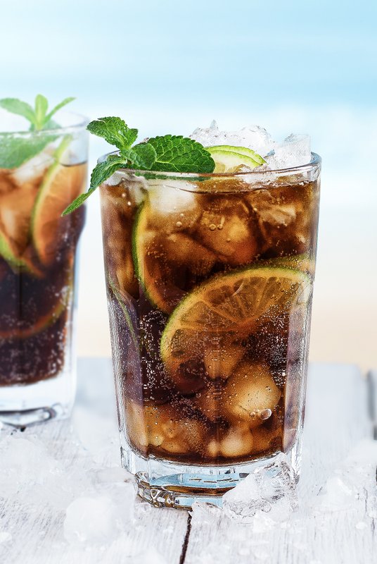 Zwei Gläser Cola mit Eis und Limetten, umgeben von Eiswürfeln, auf einem weißen Holztisch mit Meerblick im Hintergrund.