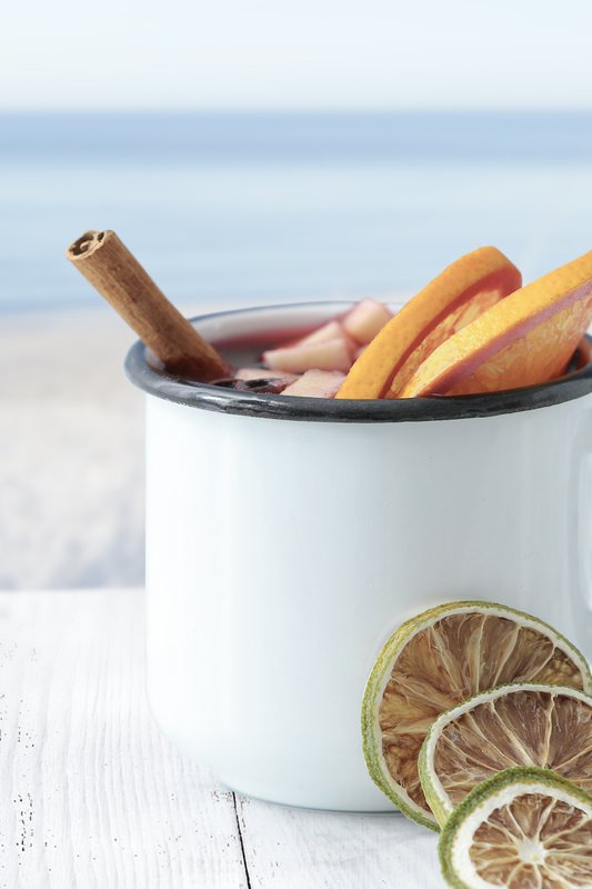 Ein weißer Becher mit Zimtstange und frischen Obstscheiben steht auf einem Holztisch, im Hintergrund der ruhige Strand.