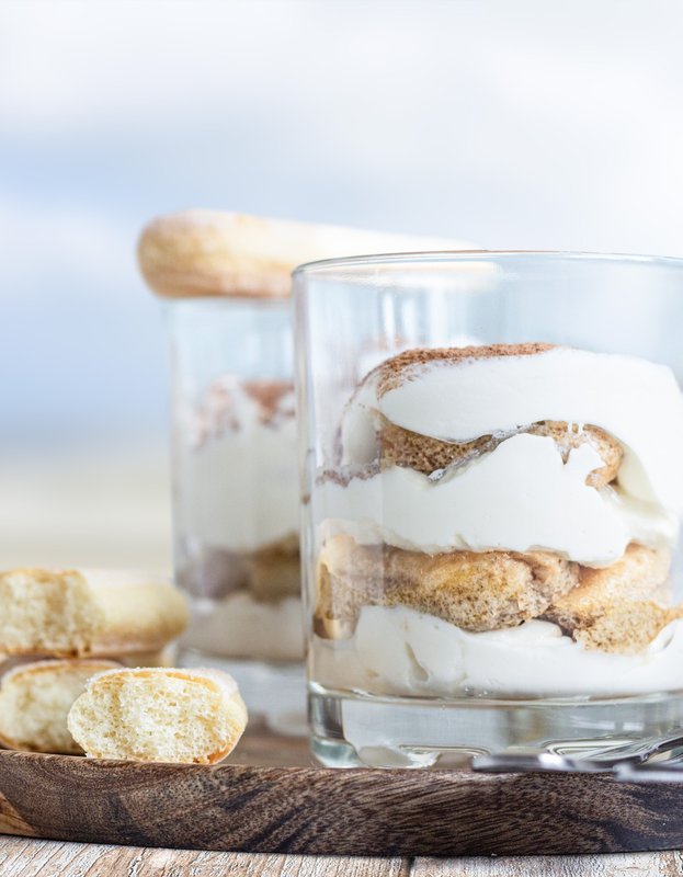 Zwei Gläser mit Schichtdessert aus Sahne und Keksen stehen auf einem Holzbrett, umgeben von einem verschwommenen Strandhintergrund.
