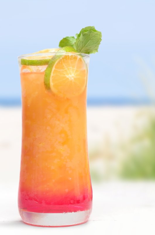 Ein erfrischendes Getränk in einem hohen Glas, gefüllt mit orangefarbenem Saft und einem roten Bodensatz, steht am Strand.