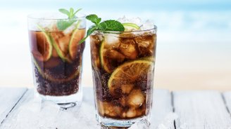 Zwei Gläser Cola mit Eis und Limetten, umgeben von Eiswürfeln, auf einem weißen Holztisch mit Meerblick im Hintergrund.