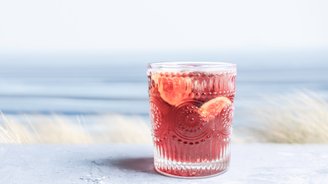 Das Glas mit dem roten Getränk steht auf einer hellen Oberfläche, während der Ozean im Hintergrund sanft im Licht schimmert.