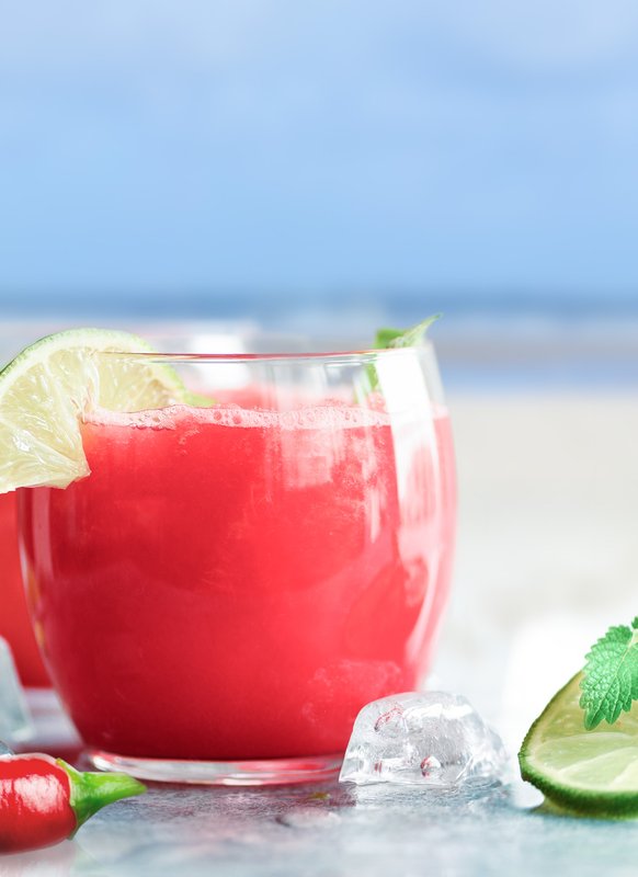 Zwei Gläser mit einem leuchtend roten Getränk, garniert mit Limettenscheiben und frischer Minze, stehen auf einem Tisch am Strand.