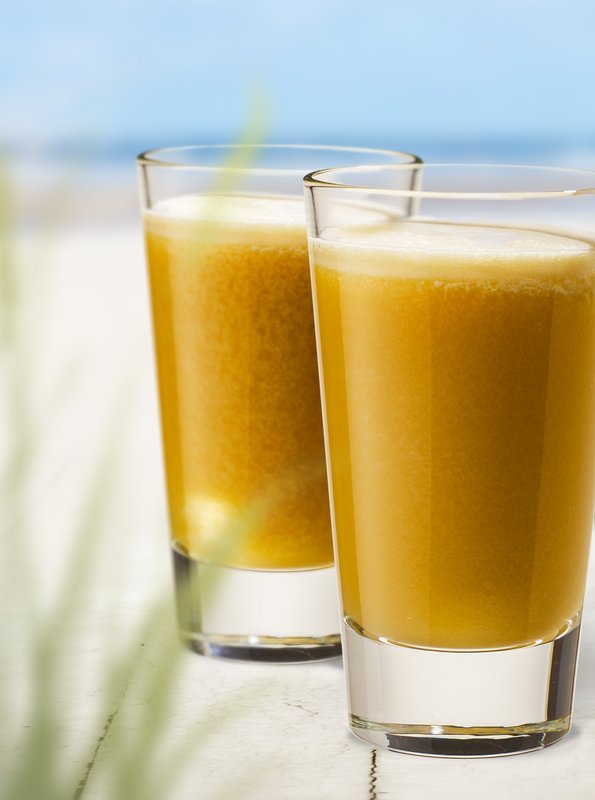 Zwei Gläser mit frischem, orangefarbenem Getränk stehen auf einem weißen Tisch, umgeben von einem Strandhintergrund.