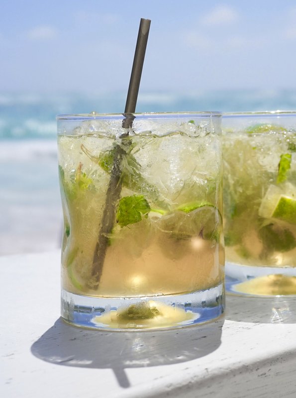 Zwei erfrischende Cocktails mit Limetten und Minze stehen auf einer weißen Holzoberfläche, während der Ozean im Hintergrund glitzert.