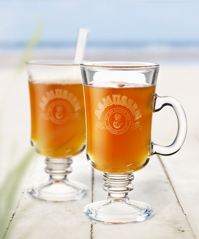 Zwei Gläser mit einem goldbraunen Getränk stehen auf einem weißen Tisch, umgeben von einem sanften Strandhintergrund.
