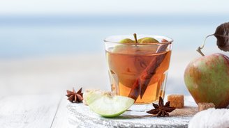 Ein Glas mit einem warmen Getränk, gefüllt mit Äpfeln und Zimt, steht auf einem rustikalen Holzbrett neben frischen Äpfeln.
