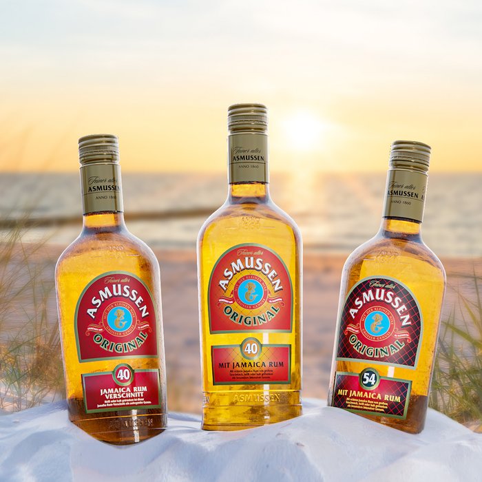 Drei Flaschen Asmussen Rum stehen auf einem weißen Sandstrand, umgeben von sanften Gräsern und dem glühenden Sonnenuntergang.
