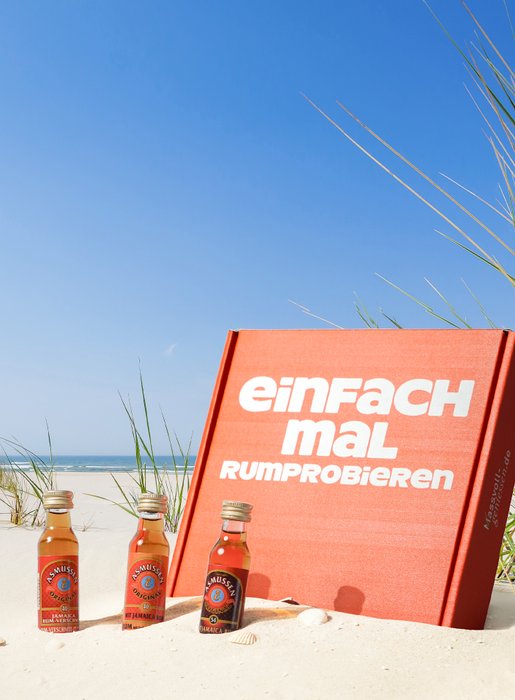 Ein leuchtend rotes Buch steht im Sand, umgeben von grünen Gräsern und drei Flaschen auf einem sonnigen Strand.