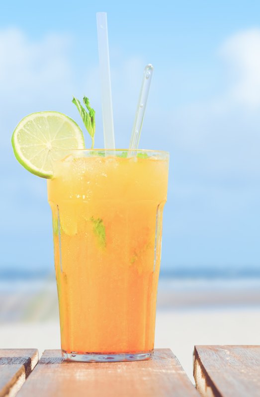 Ein erfrischendes, orangefarbenes Getränk mit Limettenscheibe und Minze steht auf einem Holztisch am Strand.