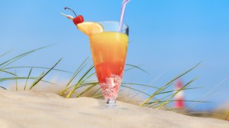 Ein erfrischender Cocktail mit einer Kirsche und einer Orangenscheibe steht auf dem Sand, umgeben von grünem Gras.