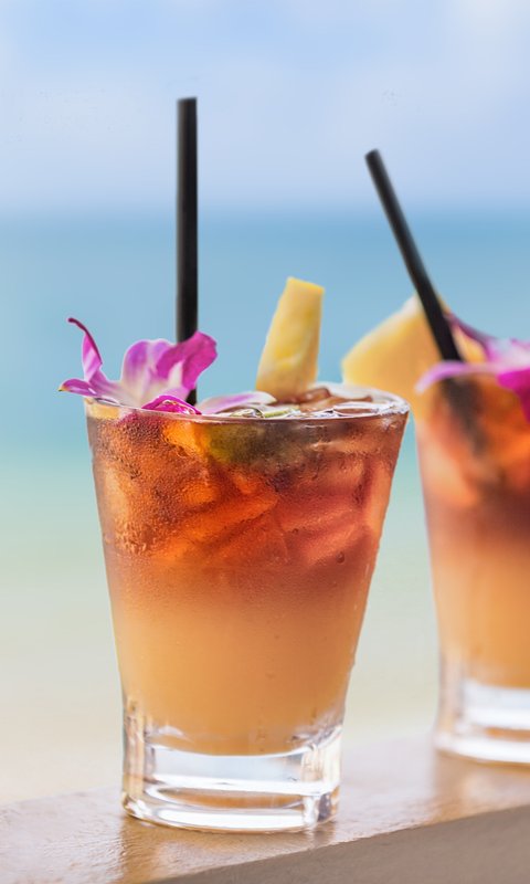 Zwei farbenfrohe Cocktails mit Eis, dekoriert mit einer Blume und Ananasstücken, stehen auf einem Tisch am Strand.