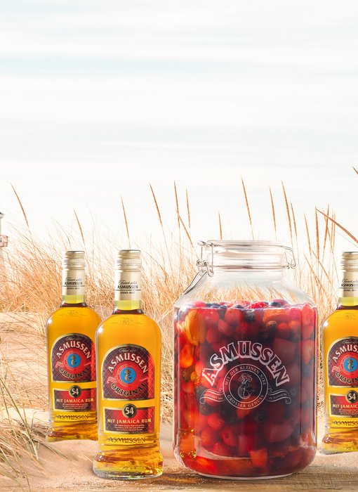Ein Glas mit bunten Früchten steht im Sand, umgeben von drei Flaschen Rum, während im Hintergrund ein Leuchtturm sichtbar ist.