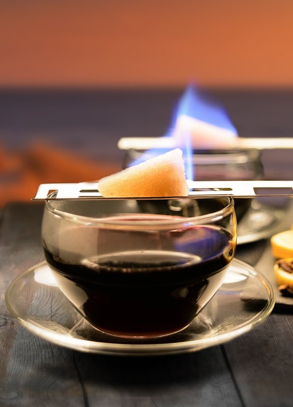 Auf einem rustikalen Tisch brennt ein Zuckerwürfel über einem Glas Kaffee, umgeben von Zimtstangen und Orangenscheiben.