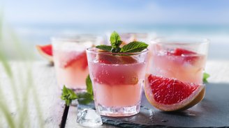 Erfrischende Cocktails in klaren Gläsern, umgeben von Eiswürfeln und Grapefruit, vor einem verschwommenen Strandhintergrund.