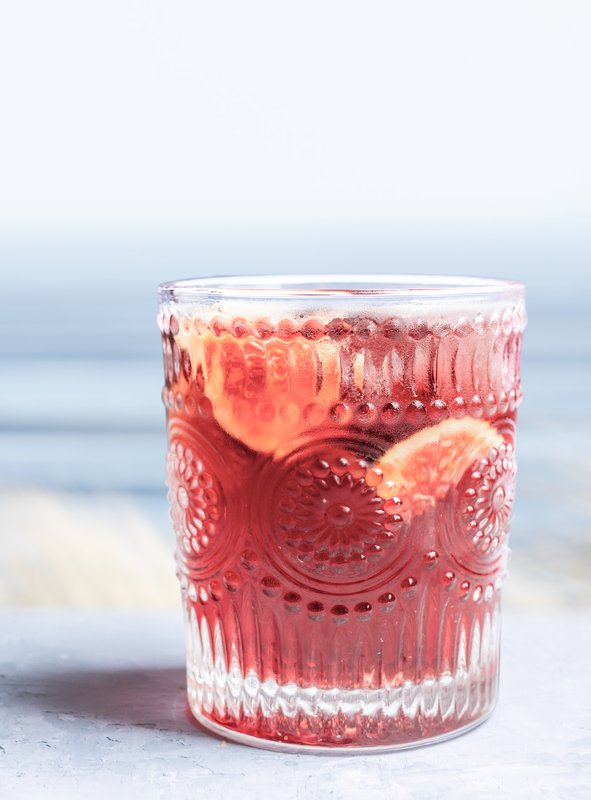 Das Glas mit dem roten Getränk steht auf einer hellen Oberfläche, während der Ozean im Hintergrund sanft im Licht schimmert.