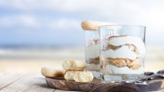 Zwei Gläser mit Schichtdessert aus Sahne und Keksen stehen auf einem Holzbrett, umgeben von einem verschwommenen Strandhintergrund.