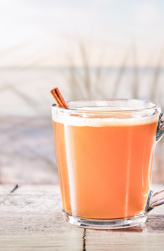 Ein Glas mit orangefarbenem Getränk steht auf einem Holztisch, im Hintergrund sind sanfte Wellen und Gras zu sehen.
