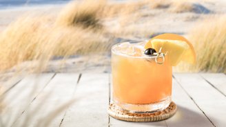 Ein köstlicher Drink in einem klaren Glas, dekoriert mit einer Ananasscheibe und einer Kirsche, vor einer idyllischen Küstenlandschaft.