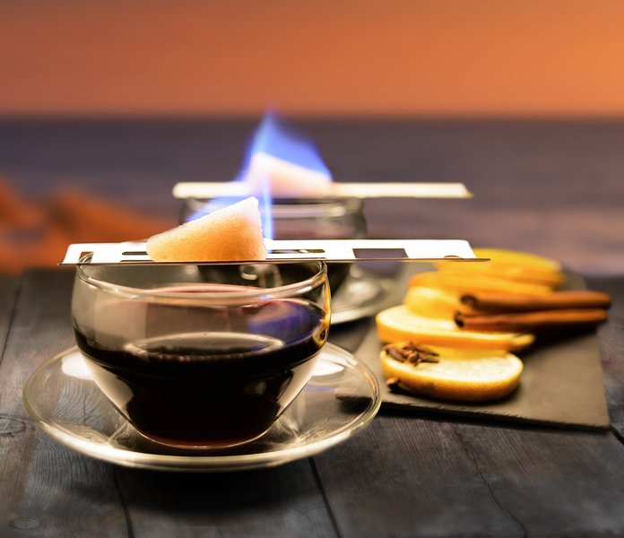 Auf einem rustikalen Tisch brennt ein Zuckerwürfel über einem Glas Kaffee, umgeben von Zimtstangen und Orangenscheiben.
