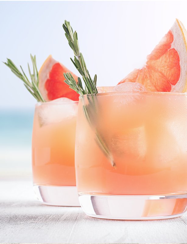 Zwei stilvolle Cocktails mit Grapefruit und Rosmarin auf einem hellen Tisch, während das Meer sanft im Hintergrund glitzert.