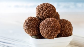 Eine Schüssel mit schokoladigen, kugelförmigen Leckereien, die mit einer knusprigen Schicht überzogen sind, steht auf einem hellen Tisch.