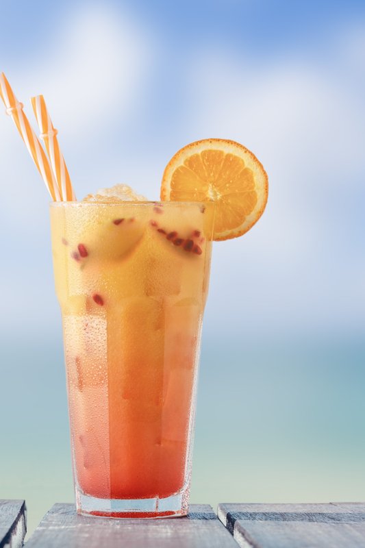 Ein buntes Cocktailglas mit orangefarbenem Getränk und zwei Strohhalmen, platziert auf einer Holzoberfläche am Strand.