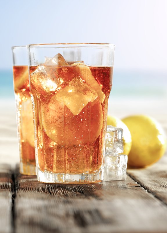 Zwei Gläser mit einem erfrischenden rötlichen Getränk, gefüllt mit Eis und Zitrone, stehen auf einem Holztisch am Strand.