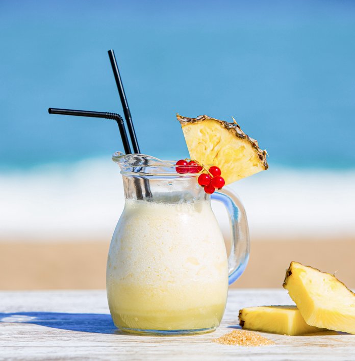 Der Krug mit cremigem Getränk steht auf einem Tisch, umgeben von frischen Ananasscheiben und einem strahlend blauen Himmel.