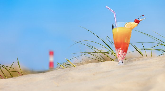 Ein farbenfroher Cocktail mit einer Orangenscheibe und einer Kirsche steht auf dem Sand, umgeben von grünem Gras.