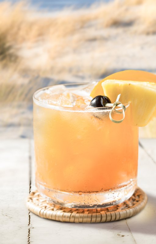 Ein köstlicher Drink in einem klaren Glas, dekoriert mit einer Ananasscheibe und einer Kirsche, vor einer idyllischen Küstenlandschaft.