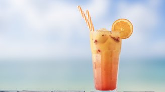 Ein buntes Cocktailglas mit orangefarbenem Getränk und zwei Strohhalmen, platziert auf einer Holzoberfläche am Strand.
