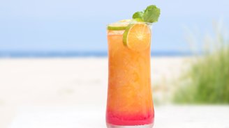 Ein erfrischendes Getränk in einem hohen Glas, gefüllt mit orangefarbenem Saft und einem roten Bodensatz, steht am Strand.