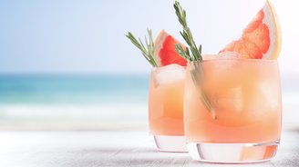 Zwei stilvolle Cocktails mit Grapefruit und Rosmarin auf einem hellen Tisch, während das Meer sanft im Hintergrund glitzert.