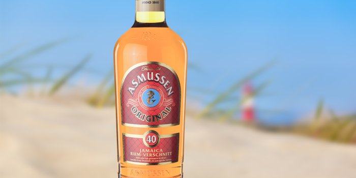 Eine Flasche Asmussen Rum steht im Vordergrund, während der Hintergrund eine verschwommene Strandlandschaft zeigt.