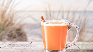 Ein Glas mit orangefarbenem Getränk steht auf einem Holztisch, im Hintergrund sind sanfte Wellen und Gras zu sehen.