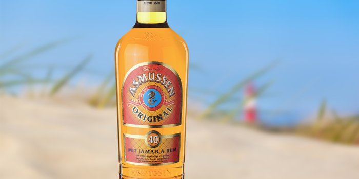 Eine Flasche Asmussen Original Rum steht vor einem verschwommenen Strandhintergrund, umgeben von sanften Gräsern.