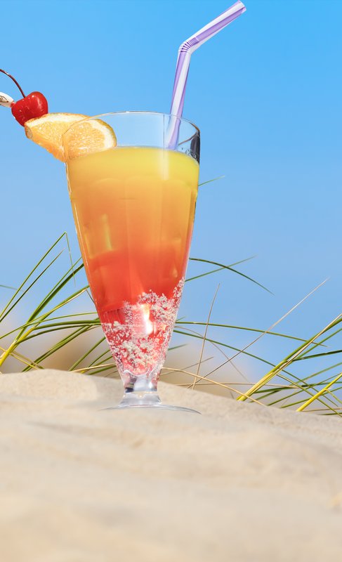 Ein farbenfroher Cocktail mit einer Kirsche und einer Orangenscheibe steht auf dem Sand, umgeben von grünem Gras.