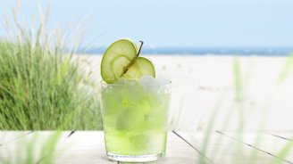 Ein Glas mit grünem Getränk und Apfelscheiben, umgeben von grünem Gras und dem blauen Meer im Hintergrund.