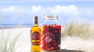 Eine Flasche Asmussen Rum steht neben einem großen Glas mit bunten Früchten, vor einer malerischen Strandkulisse.