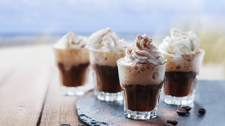 Vier kleine Gläser mit schaumigem Kaffee, gekrönt von Sahne und Schokoladenstreuseln, stehen auf einem rustikalen Tisch.