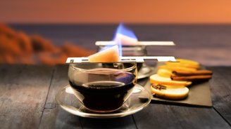 Auf einem rustikalen Tisch brennt ein Zuckerwürfel über einem Glas Kaffee, umgeben von Zimtstangen und Orangenscheiben.