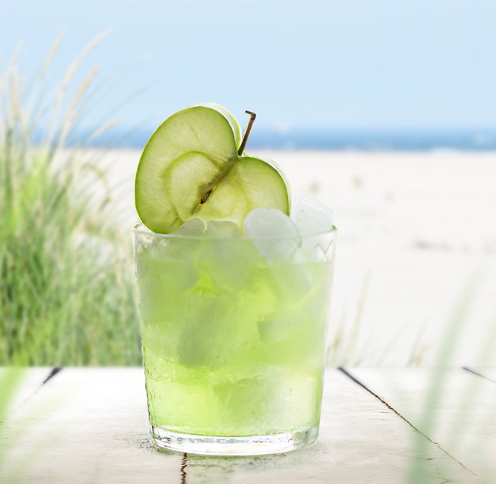 Ein Glas mit grünem Getränk und Apfelscheiben, umgeben von grünem Gras und dem blauen Meer im Hintergrund.